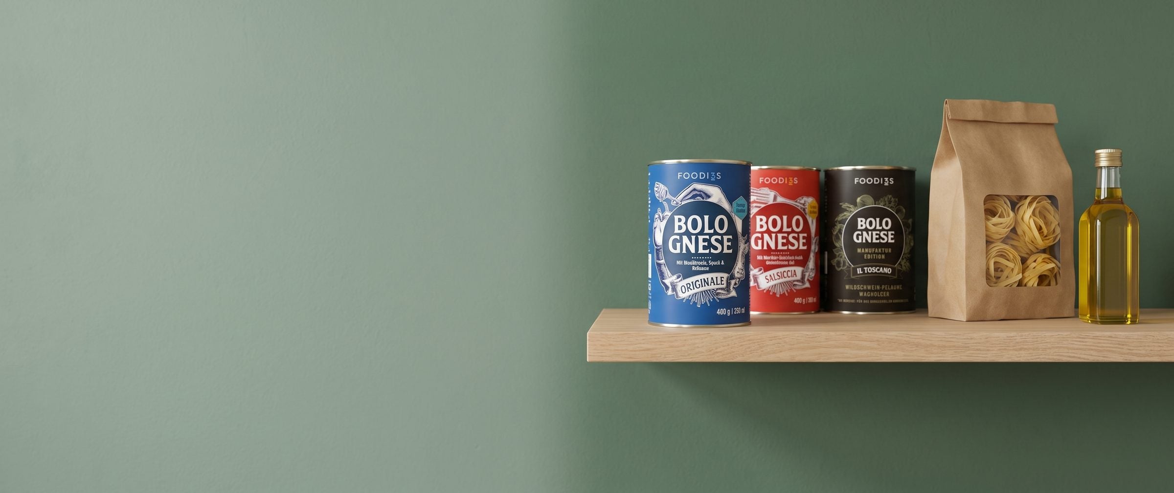 Ein Regalbrett aus Eiche an einer salbeigrünen Küchenwand, darauf Dosen 3foodies Bolognese Originale, Salsiccia und Il Toscano, eine Tüte Eiernudeln und eine Flasche Olivenöl